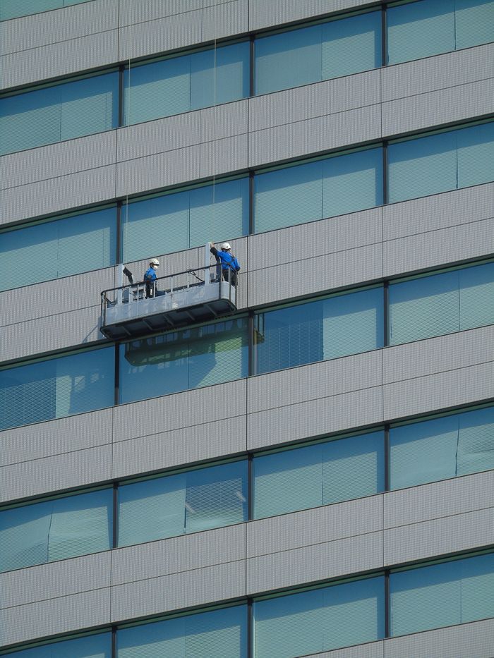 our-story Two workers clean windows on a modern glass building facade using a suspended platform.