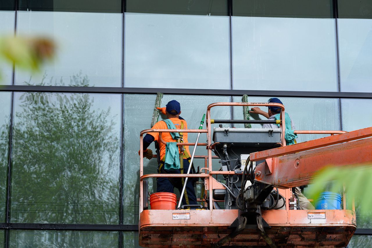about-img-01 Two workers cleaning windows on a glass building facade using a lift platform, enhancing building aesthetics.